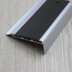 A black and silver aluminum strip, also known as the Warrior Stair Nosing (ASN5020R), placed on a wooden surface with a rubber insert.