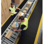 Two men, wearing Warrior Rubber Anti Fatigue ESD Mats 60CM Width 90CM Length Black and Yellow WAFM-9060-B/Y, are working on a conveyor belt in a warehouse.